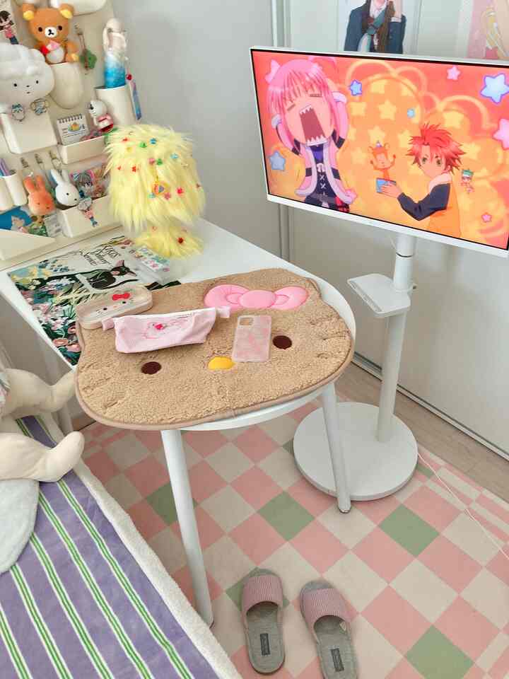 白とピンクの色調の寝室、アニメが流れるテレビとかわいいキャラクターの小物が配置された雰囲気のあるオタク風インテリア