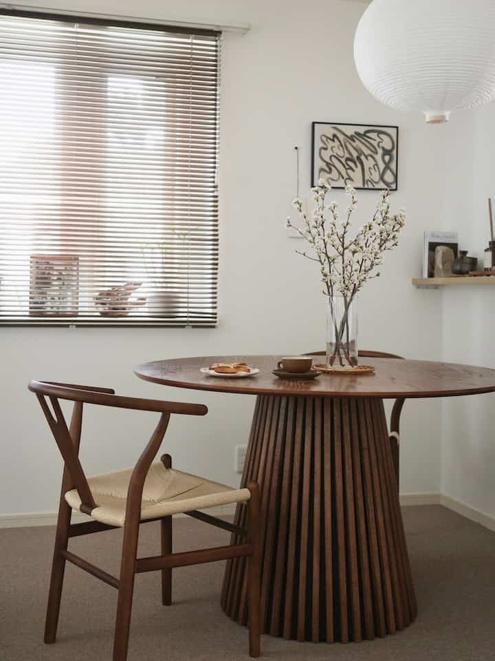 Warm wood tone dining room with white walls, featuring a round wooden dining table and chairs with a vase of blossoms, minimalistic and natural ambiance