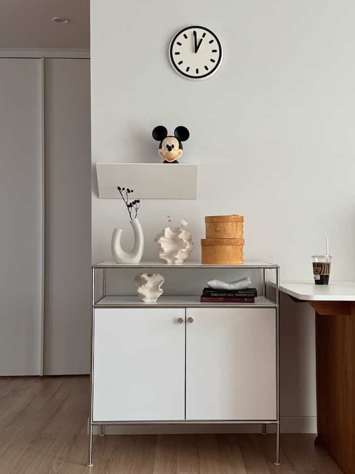 Natural wood and white toned living room featuring a minimalist cabinet, sculptural objects, and a wall clock creating a modern atmosphere