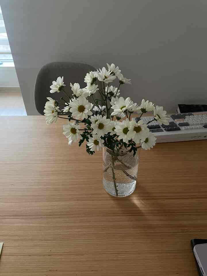 ナチュラルな木目調のテーブルに白い菊の花が入った透明な花瓶とグレーのダイニングチェアが配置されたシンプルなダイニングルーム