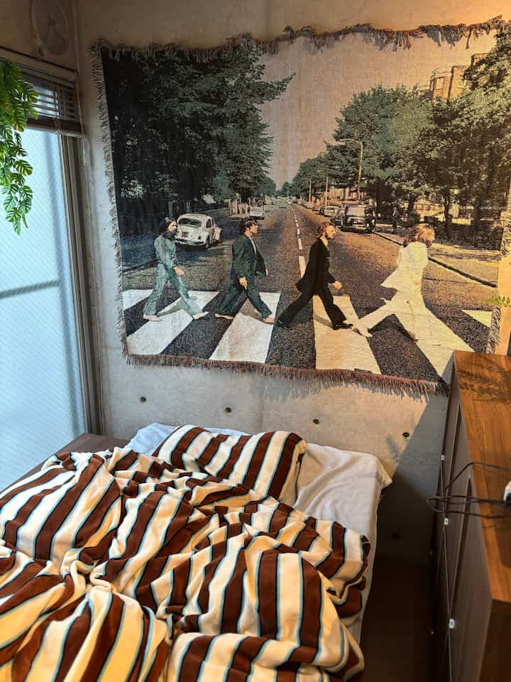 ベージュとブラウンの色調の寝室、ビートルズの壁掛け布とストライプのブランケットが置かれた印象的な空間