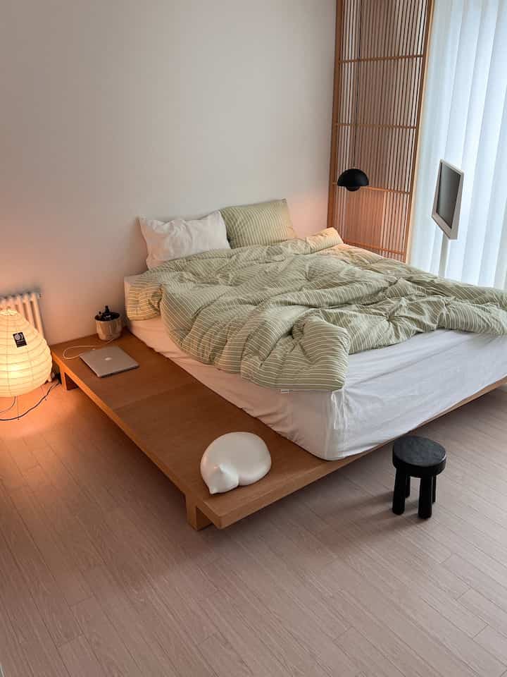 白と木目調が調和したシンプルでモダンな寝室、中央にストライプの寝具が置かれたローベッドと木製のパーティションが配置された印象的な空間