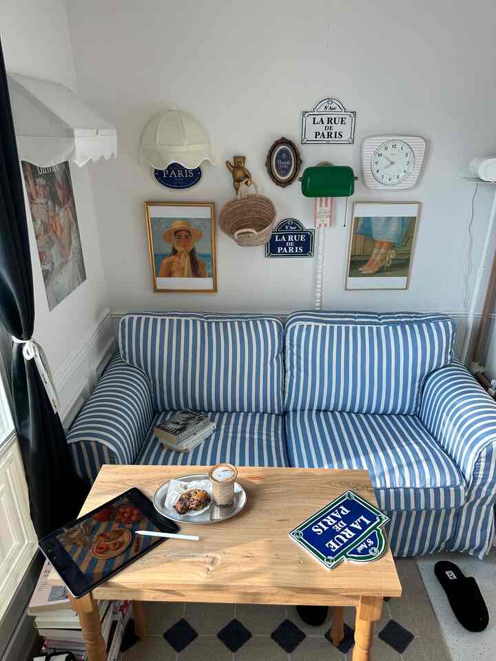 白と青のトーンの小さなリビング、ストライプのソファと木製コーヒーテーブル、フレンチヴィンテージの壁飾りが調和した雰囲気のあるインテリア