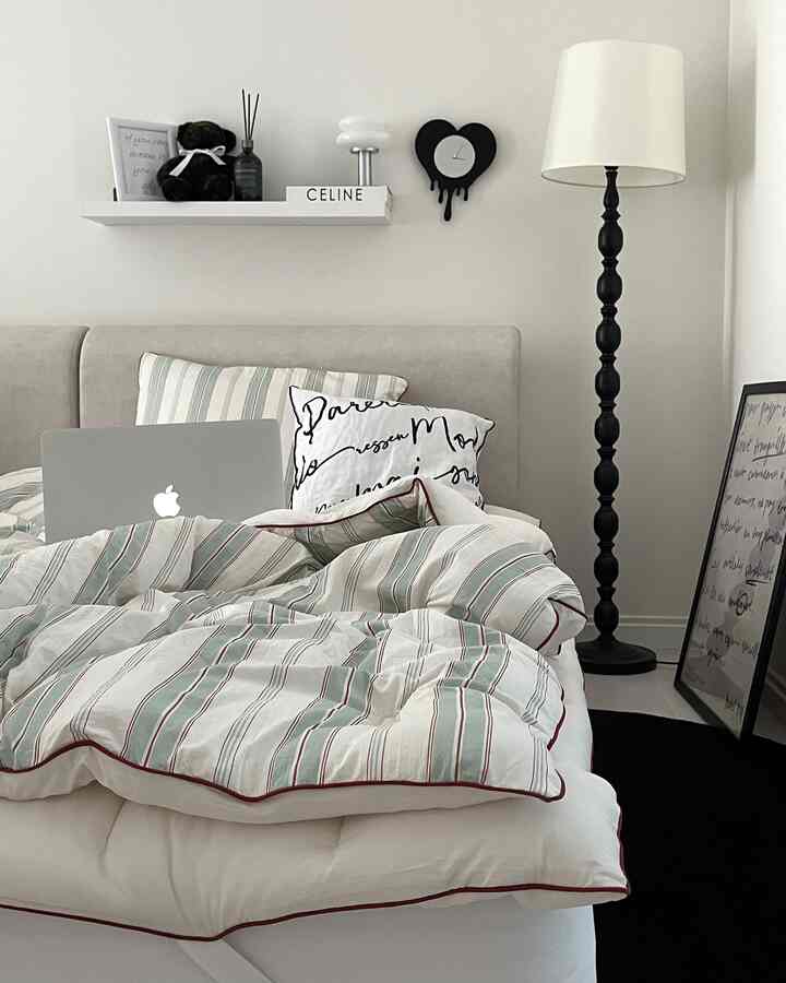 A white and gray toned bedroom featuring a bed with cushions centrally placed, a floor lamp and a poster on the right side creating a clean space