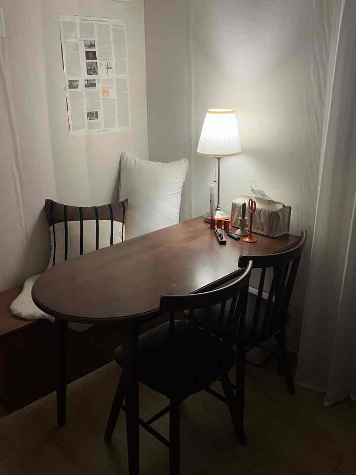 ライトブラウンの木製丸テーブルと椅子、コーナーベンチ、白い壁と照明が配されたワンルームのキッチンスペース