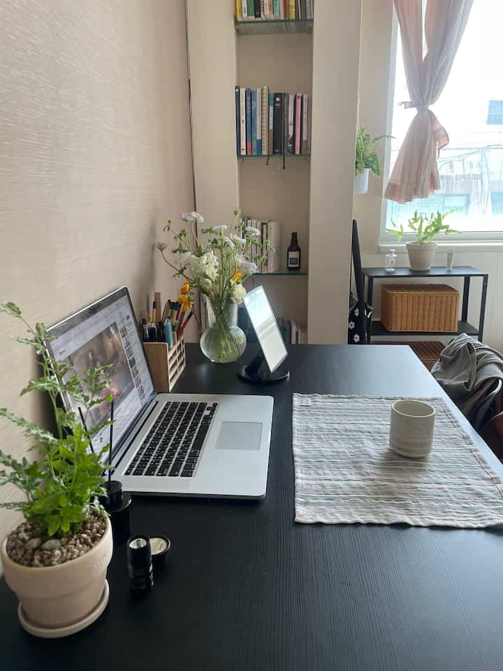 白い壁と黒いデスクが特徴のワンルーム作業スペース、観葉植物と窓辺のカーテンで落ち着いた雰囲気のインテリア