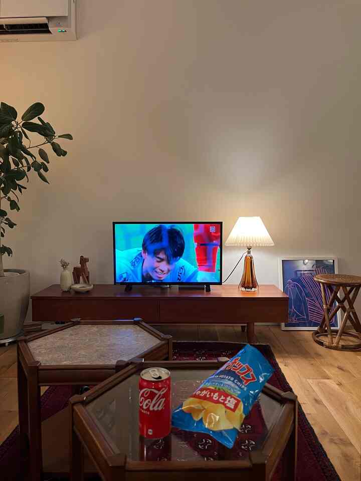 ナチュラルなトーンの木製家具とテーブルランプが配置されたリビング、テレビとスナックが置かれたくつろげる空間