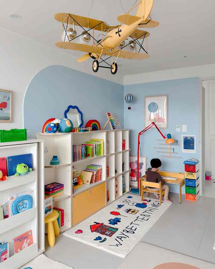 淡いブルーの壁と白い家具、黄色い木製の机とカラフルな収納ボックスが配置された子供部屋で、子供が机に座っている様子