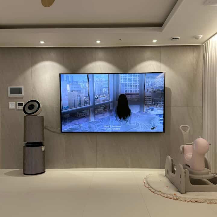 ベージュの壁と白いタイルの床があるリビング、壁掛けテレビと子供用おもちゃが配置された清潔感のある空間
