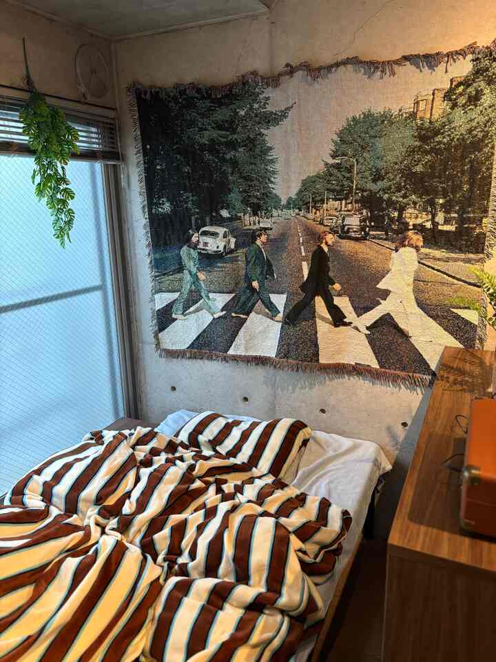 ベージュと木目調が調和した一人暮らし用の居心地の良い寝室、ビートルズのタペストリーとストライプのブランケットが印象的なインテリア