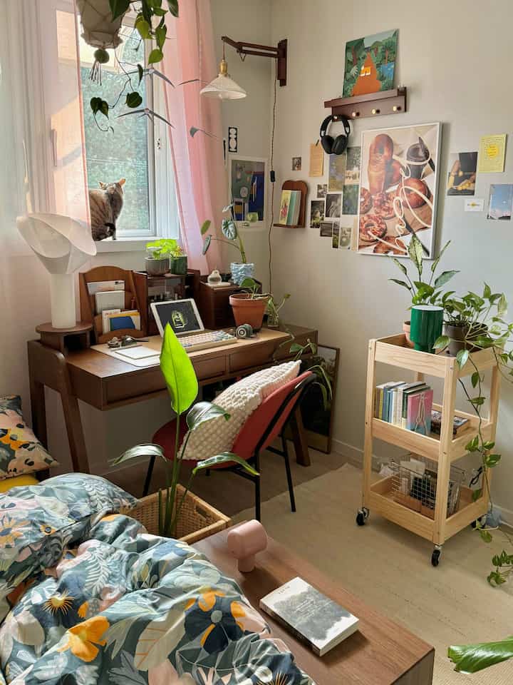 ナチュラルな木目調とピンクのカーテンが調和した寝室兼ホームオフィス、窓辺に座る猫と植物が配置された雰囲気のあるインテリア