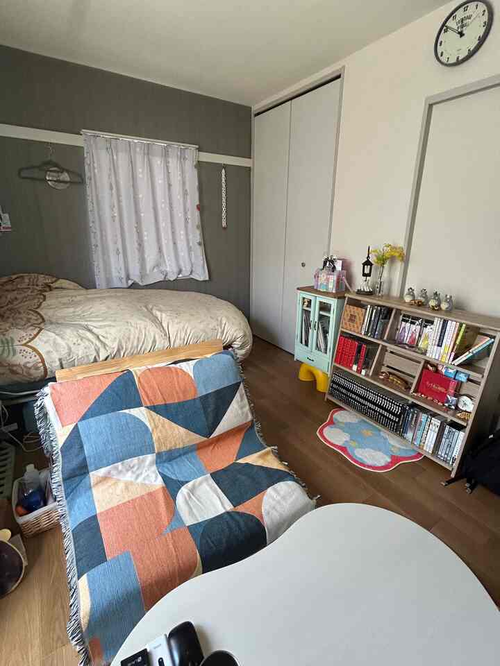 A cozy 1K studio bedroom with white and gray walls, featuring a bed, low sofa with patterned cover, and bookshelf.