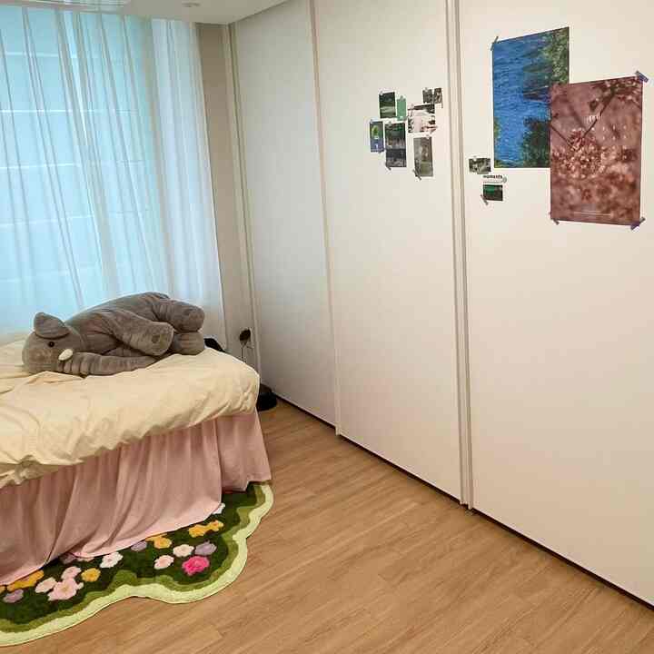 白い壁と木目調の床が調和する一人暮らしの寝室、ピンクのベッドカバーと花柄のラグが温かみのあるインテリア