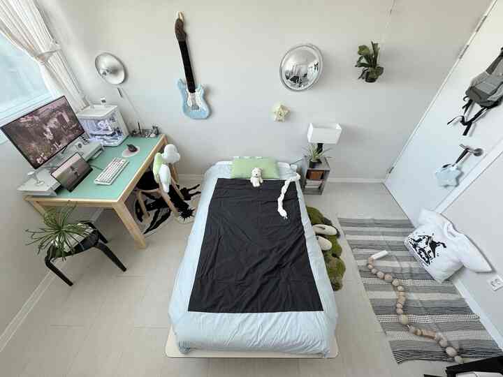 明るい木目調のデスクと黒の掛け布団カバーがある小さなワンルーム寝室、モニターと植物が配置された整ったホームオフィス