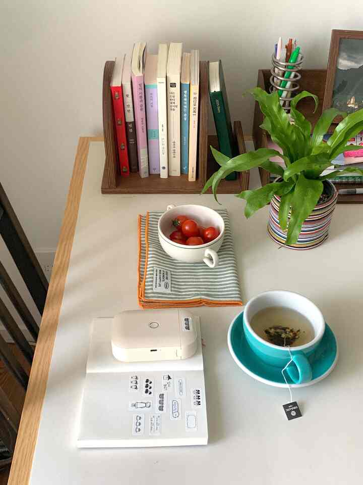 白いデスクの上に木目調の本立て、植物、ティーカップ、トマトが配置された落ち着いたホームオフィス空間