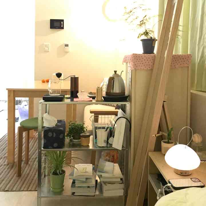 ナチュラルな色調のワンルーム、木製デスクと棚、植物、照明が配置された雰囲気のあるホームカフェ風インテリア