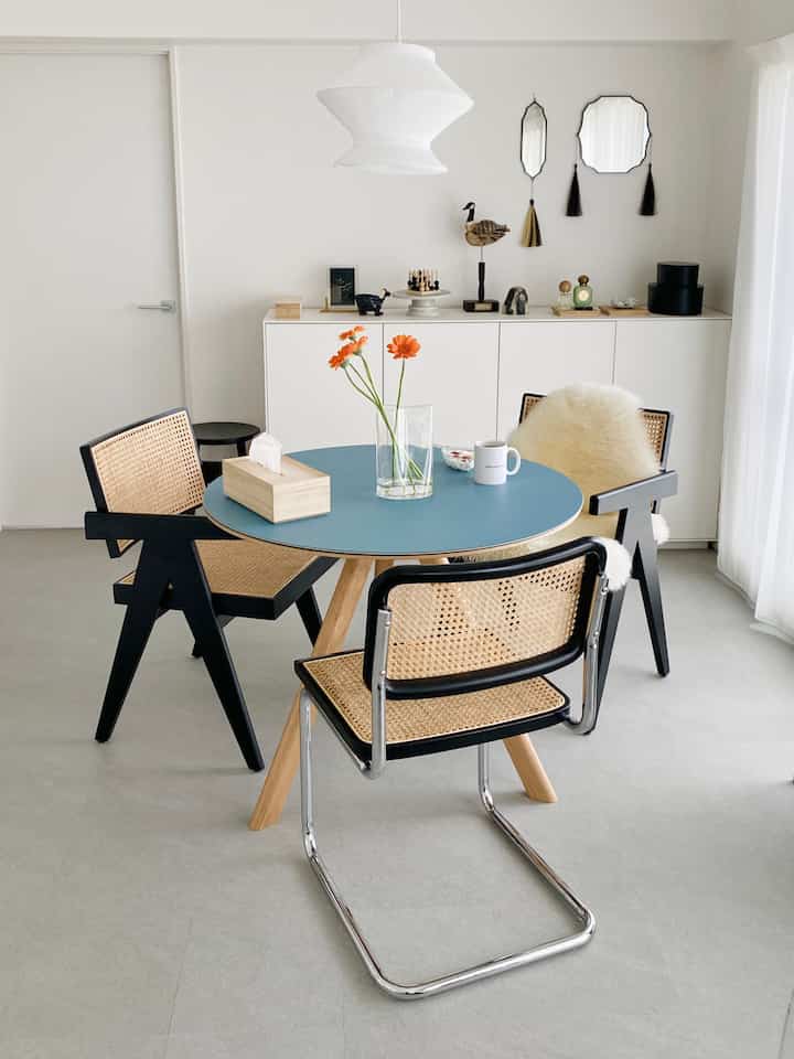 白とナチュラルな色調のダイニング、スカイブルーのテーブルマットとオレンジの花が置かれた丸いダイニングテーブルと椅子のある雰囲気のあるインテリア