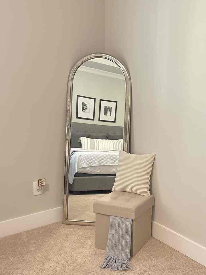 Beige-toned bedroom corner space featuring a standing mirror and foldable ottoman with a soft throw and cushion, creating a simple and cozy atmosphere