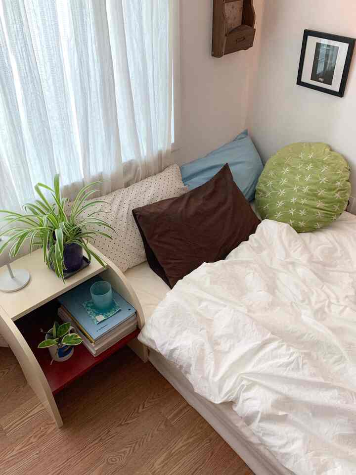 白とナチュラルトーンの小さな寝室、ベッドとクッション、ナイトテーブル、植物が配置された落ち着いたシンプルなインテリア