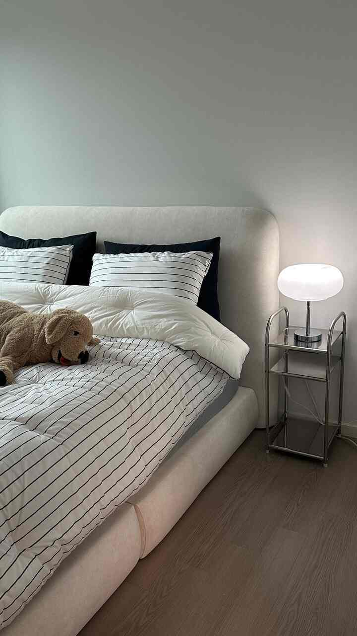 White and beige toned bedroom featuring striped bedding, stainless steel side table, and a mood lamp in a clean space