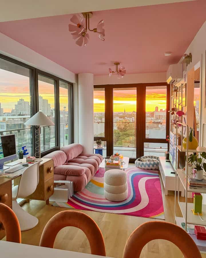 A modern living room with a pink ceiling and soft pastel-toned rug, large windows showcase a warm sunset outside.