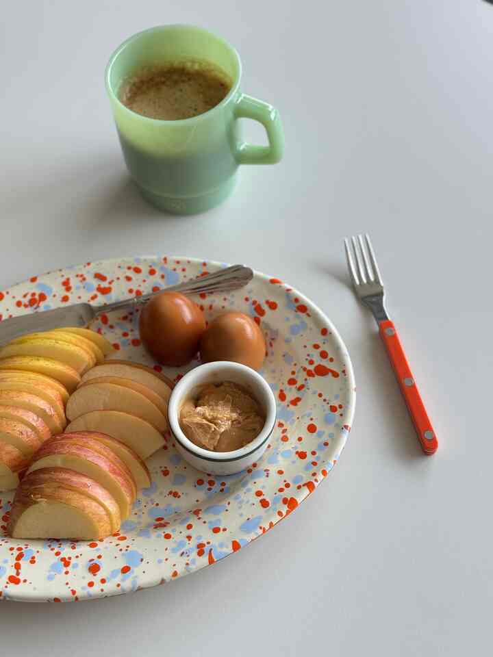 A cozy home cafe setup featuring an oval plate with orange and sky blue splatter pattern alongside a light green mug on a clean white surface