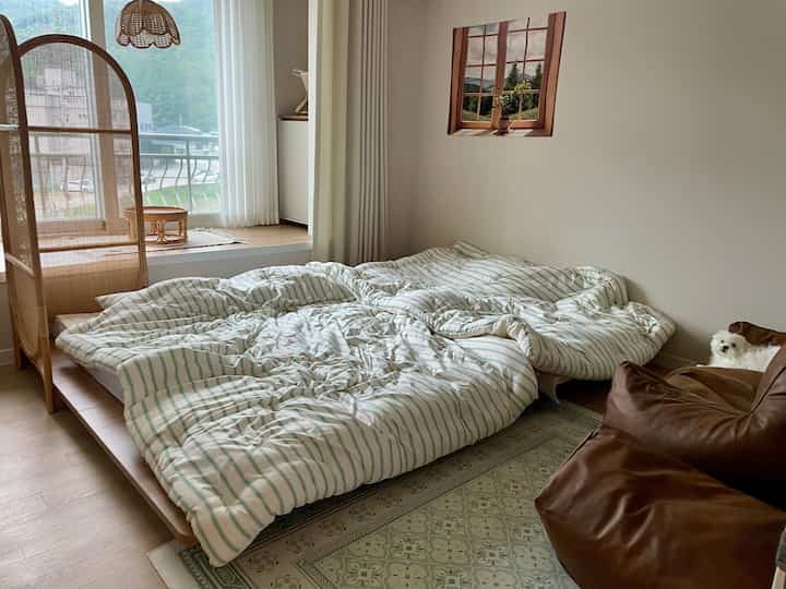 Natural beige-toned bedroom featuring low wooden bed, brown leather armchair, and a pet dog creating a cozy atmosphere