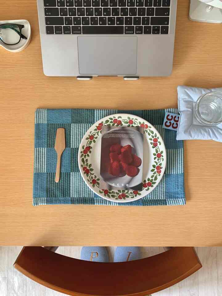 木目調のダイニングテーブルにノートパソコン、青いチェックのランチョンマット、花柄の皿とクッションが置かれた整ったホームオフィス空間