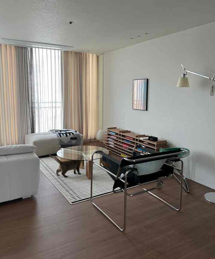 ホワイトと木目調のナチュラルモダンなリビング、中央に透明なガラステーブルと黒いフレームの椅子が配置された落ち着いた空間