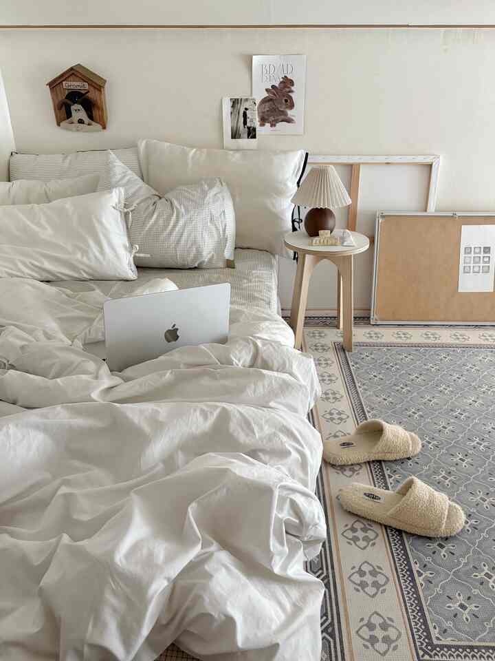 ナチュラルな色調と白い寝具が配置された寝室、木製のスツールとテーブルランプが温かみのある雰囲気を演出