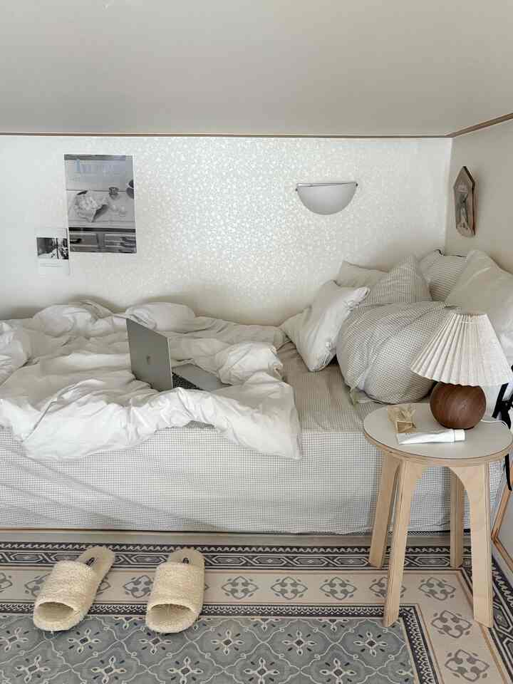 Bright white and light brown wood tone bedroom featuring clean bedding and a table lamp on a stool creating a cozy atmosphere