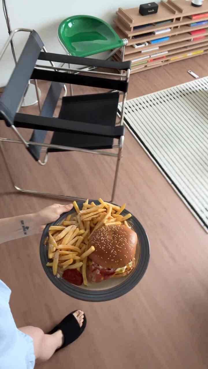 ウッドトーンの床とミッドセンチュリーモダンの椅子があるコンピュータールームで、ハンバーガーとフライドポテトの載った皿を手に持つ様子