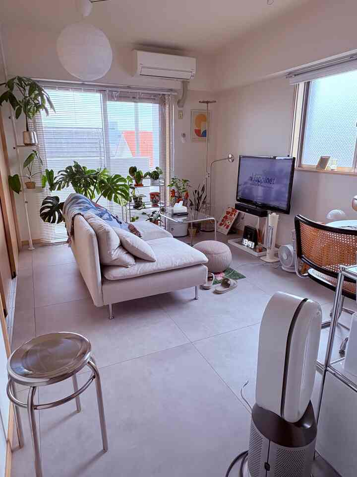Bright gray-toned living room featuring cream sofa, transparent side table, and multiple plants arranged near window for a cozy atmosphere