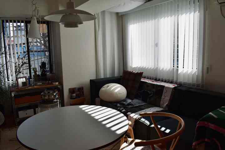 ホワイトとブラウンのトーンのリビング、窓に設置されたバーチカルブラインドとラウンドテーブル、ウィッシュボーンチェアが配置された雰囲気のある空間