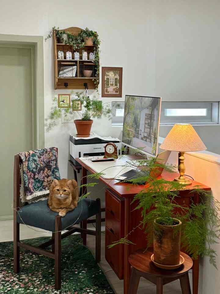 ブラウンの木目調と緑の植物が調和した小さなワークスペース、ヴィンテージのテーブルランプと猫がいる雰囲気のあるインテリア