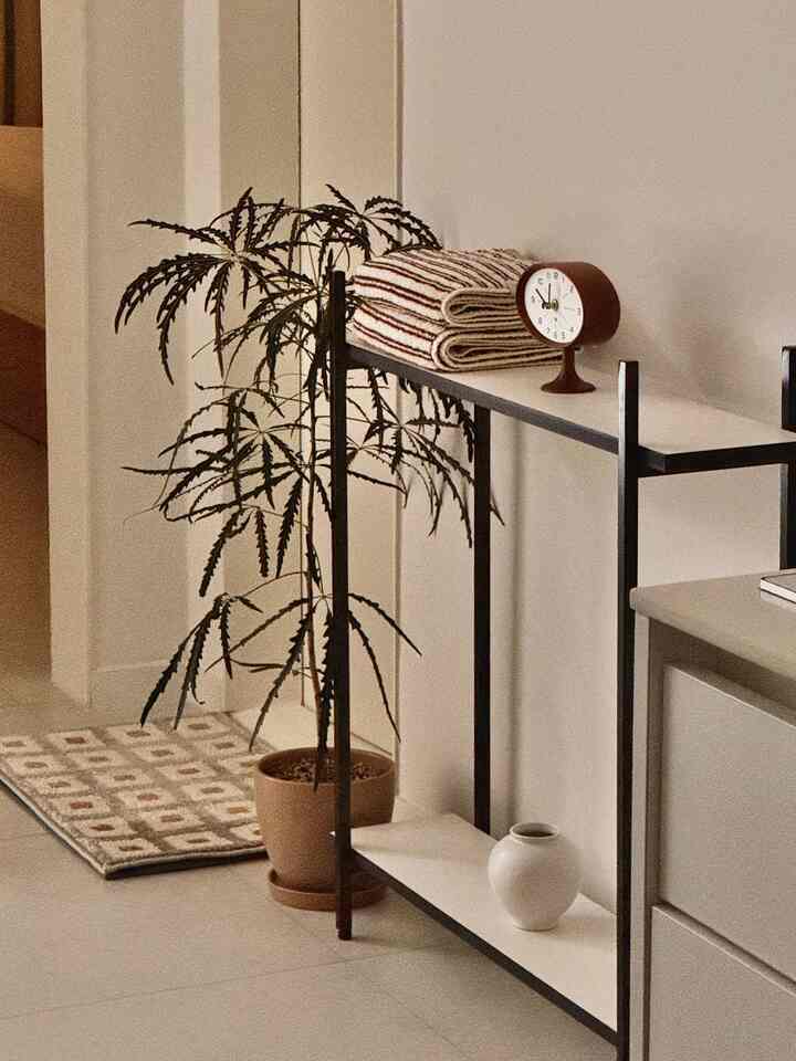 白い壁と床、黒いフレームのサイドテーブルに茶色のタオルと時計が置かれ、玄関近くにドアマットが配置されたシンプルな玄関スペース