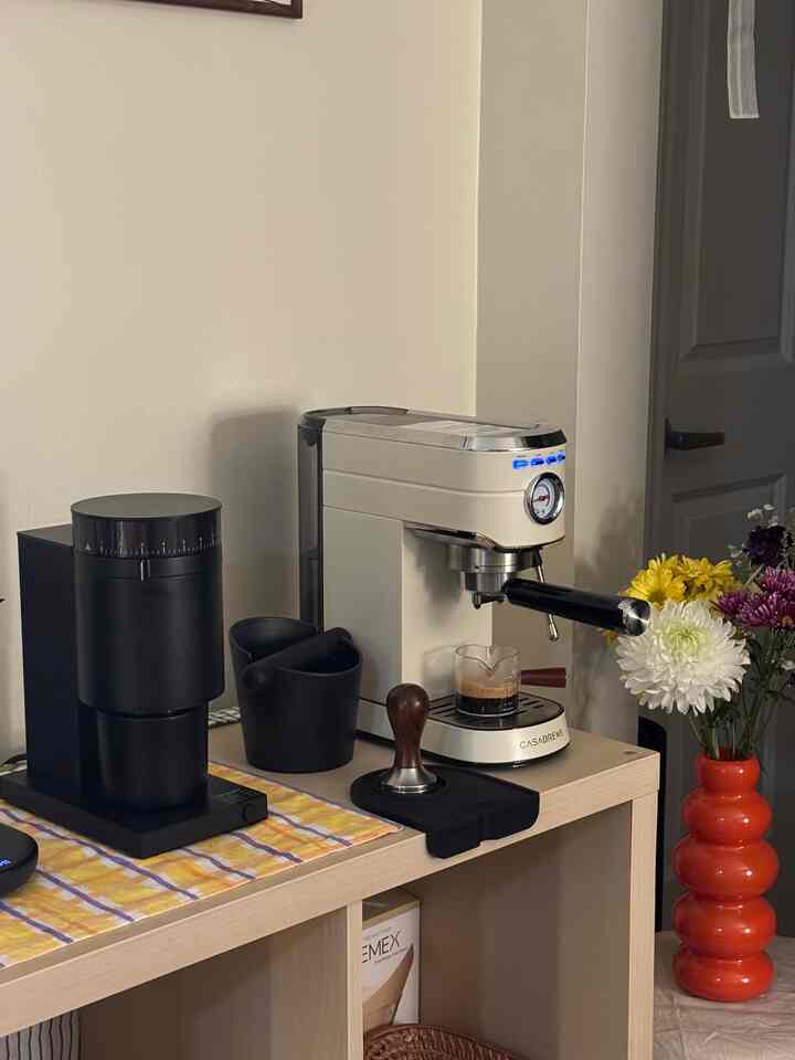 Compact home cafe space in white and black tones featuring a coffee machine, grinder, and orange vase with flowers, creating a cozy atmosphere