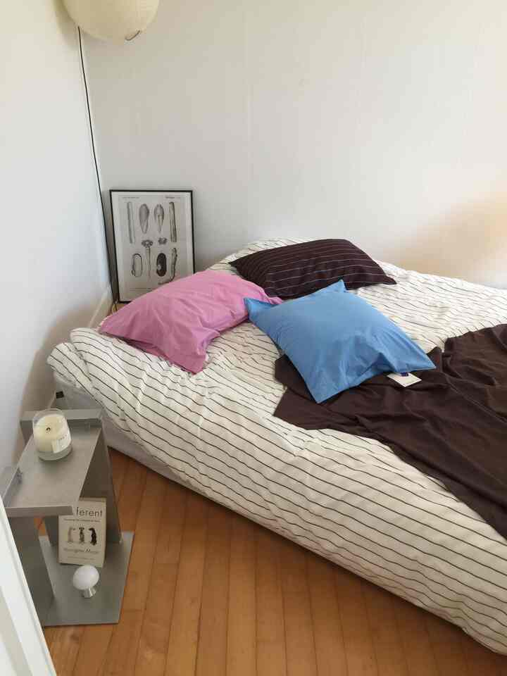 白い壁と木製の床があるシンプルな寝室、ストライプ柄の掛け布団カバーとカラフルな枕が置かれたベッドが印象的な空間