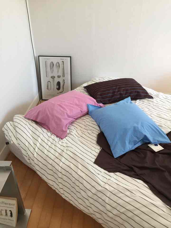 白い壁とストライプ柄の寝具があるシンプルな寝室、ピンク、青、茶色の枕が配置された雰囲気のある空間