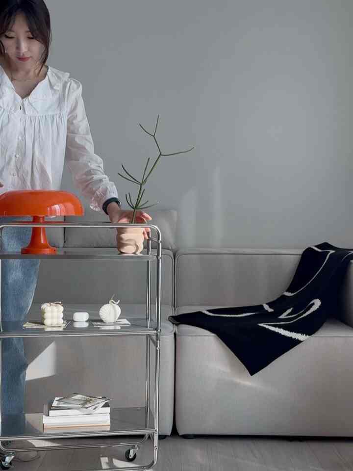 Gray-toned modern living room featuring a slim stainless steel trolley with an orange mushroom lamp, gray sofa, and black patterned throw for a simple ambiance