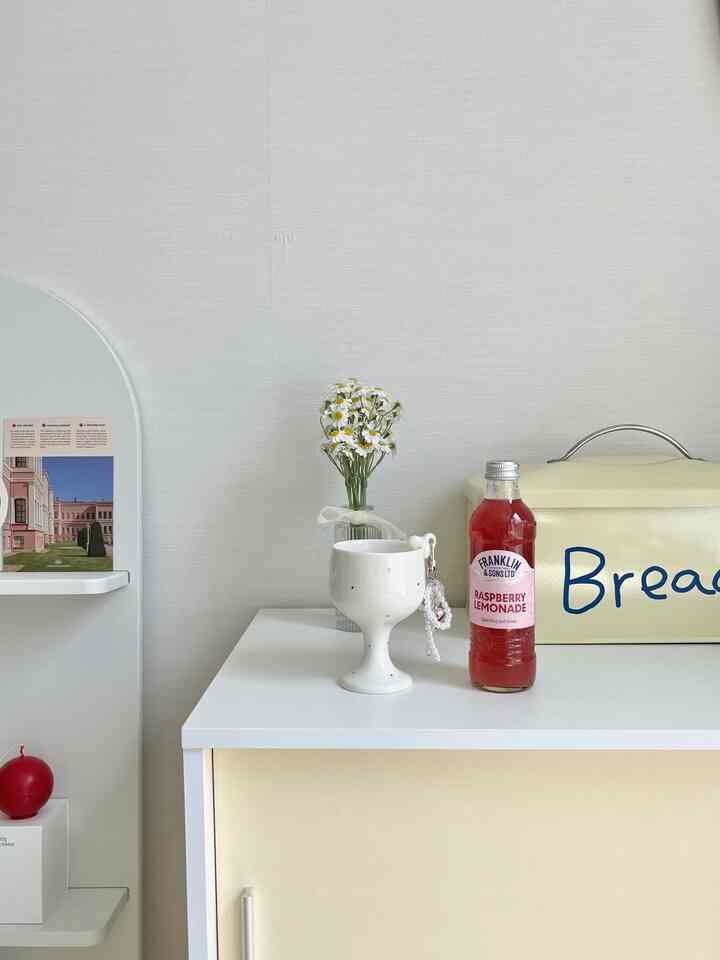 Bright white and beige toned studio kitchen space featuring ceramic goblet cup with ribbon keyring, red drink bottle, and bread box in a minimal setup