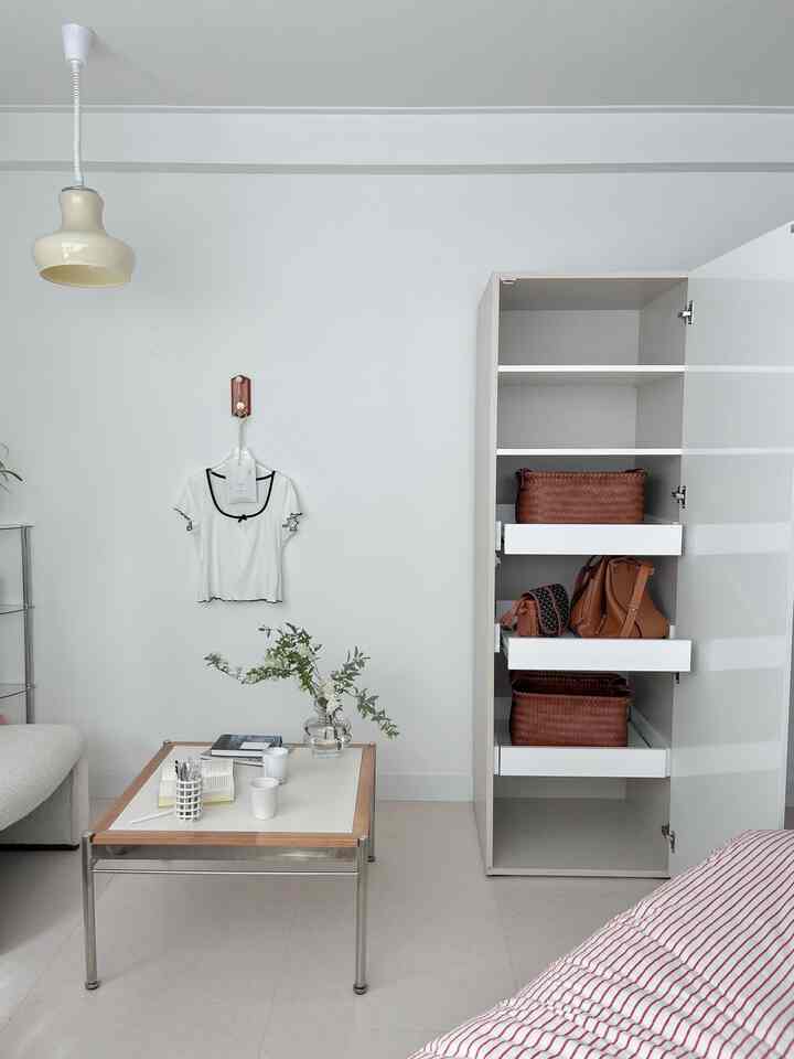 Bright white-toned bedroom featuring a simple table with plants and cups centrally, and an open white cabinet on the right, creating a tidy space