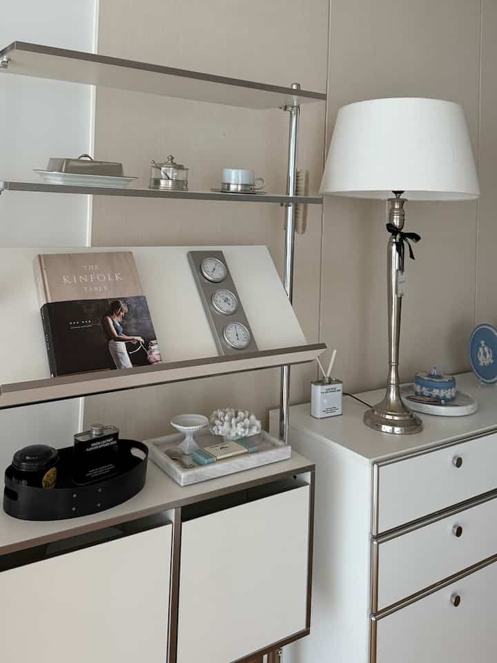 白とベージュのトーンのリビングルーム、ドレッサーと金属製の棚、ディフューザーと照明が配置された清潔感のあるインテリア