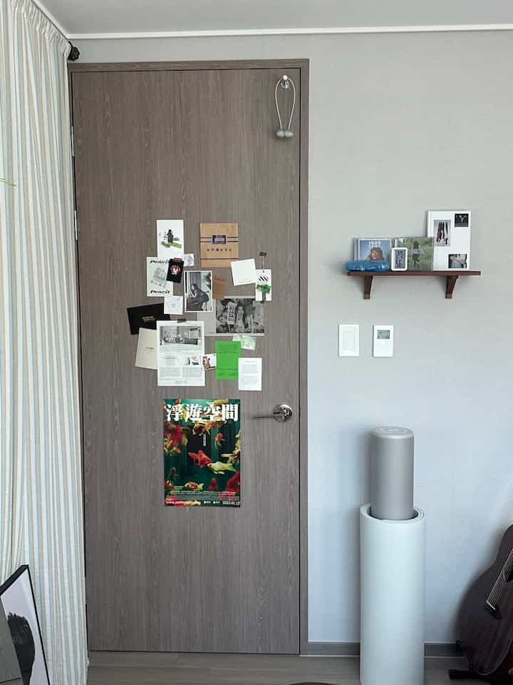 ナチュラルな木目調のドアとグレーの壁、ポスターと棚が配置された清潔感のあるワンルームの空間