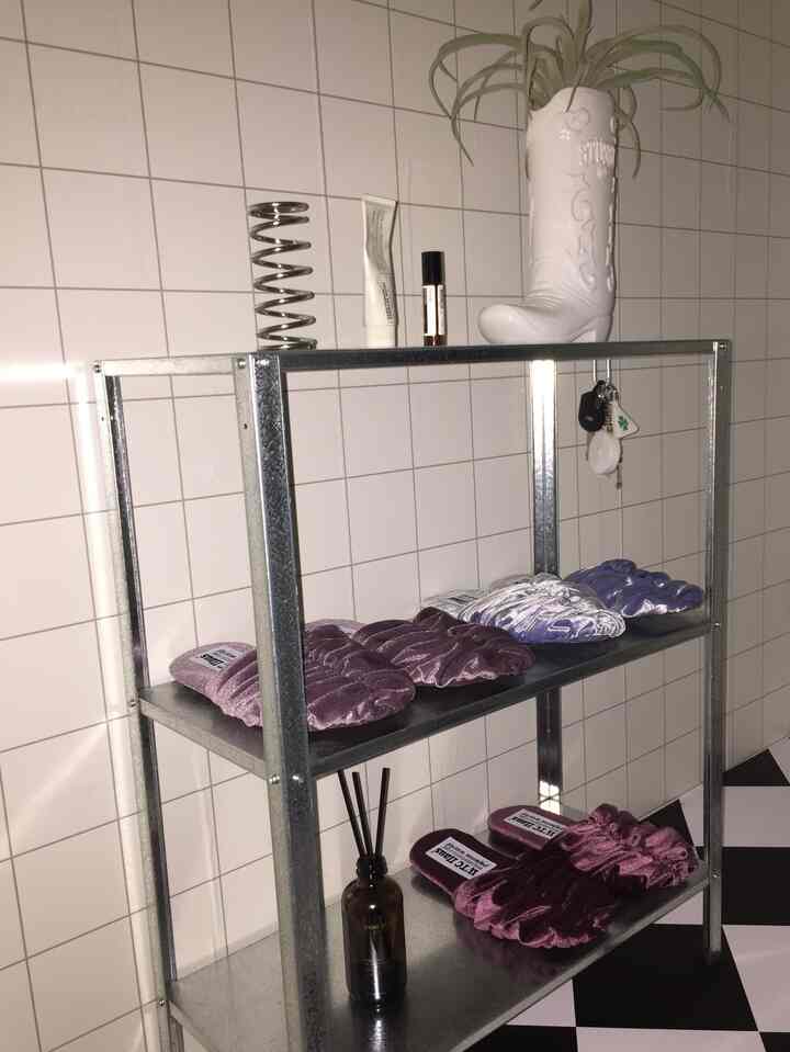 白いタイルの壁とグレーの鉄製棚があるモダンな室内、紫のスリッパと観葉植物、ディフューザーが整然と並べられたインテリア