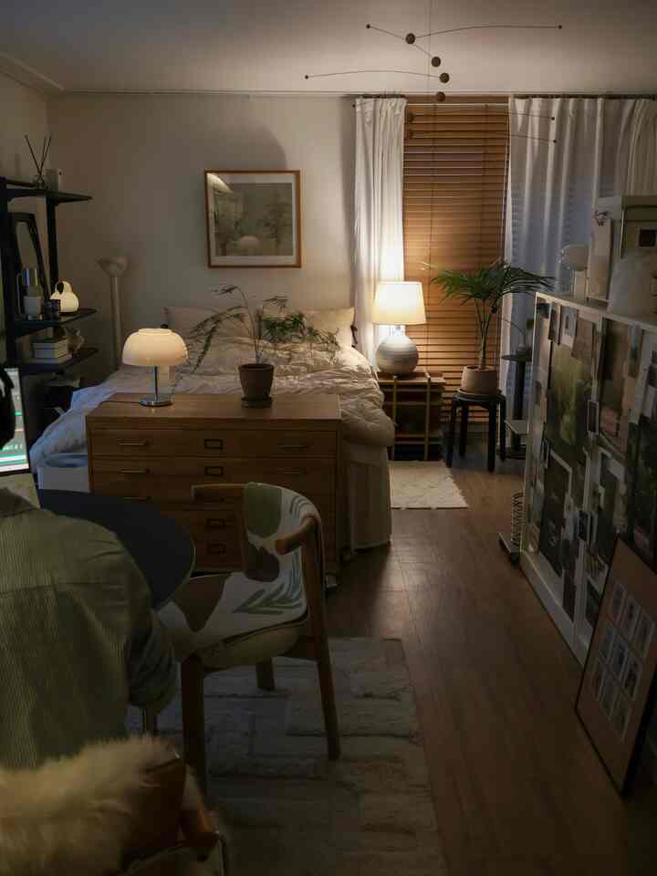 温かみのある木目調と白いカーテンが配置されたワンルーム寝室、チェストと照明が印象的な空間を演出している