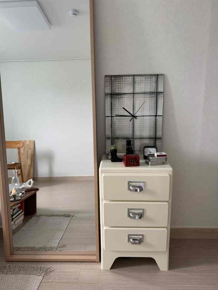 ホワイトとブラウンの色調の寝室、クリーム色のキャビネットと全身鏡、壁にはガラスブロックの時計が配置された清潔感のあるインテリア