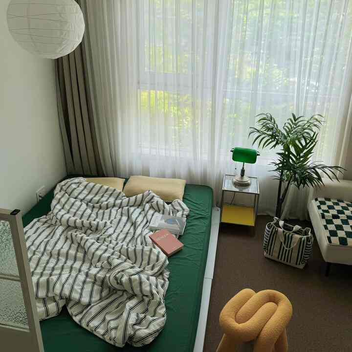Green and beige toned studio bedroom featuring low bed with striped summer bedding, plants, and mid-century modern decor in a cozy setting