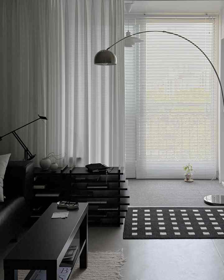 白と黒のトーンのモダンなリビングルーム、シフォンカーテンとアルミブラインド、黒いラグ、シルバーのアーチ型フロアランプが配置された印象的な空間