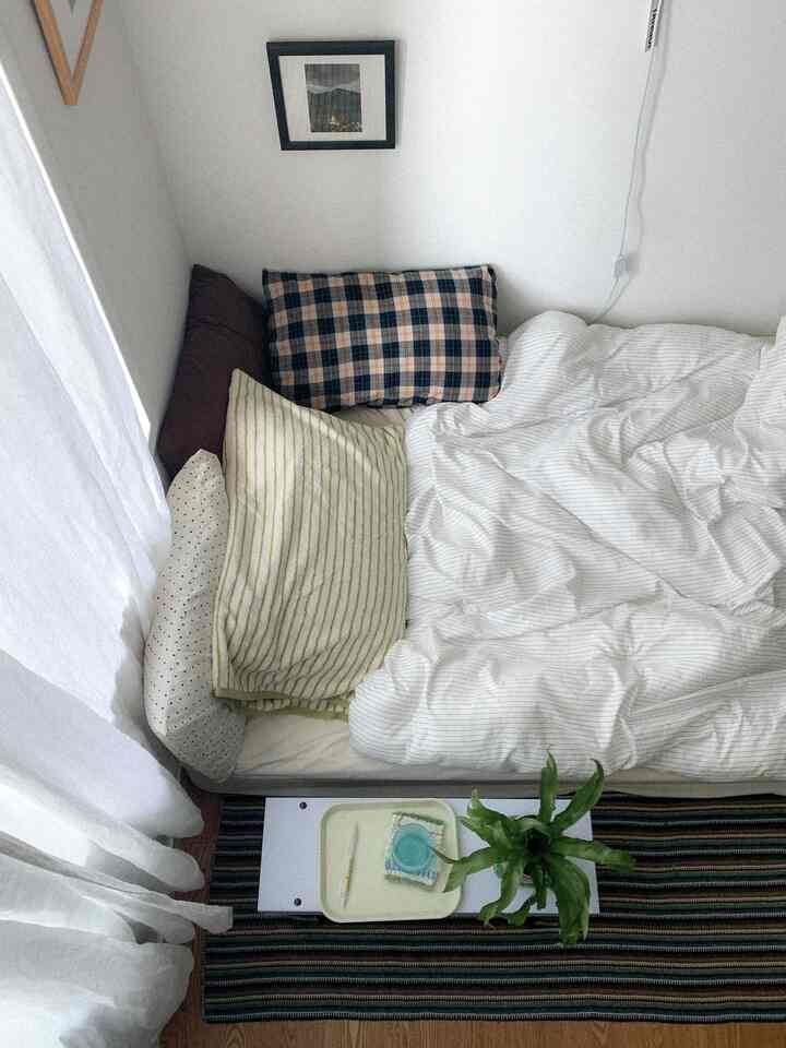 ホワイトとナチュラルトーンの一人暮らし寝室、整った寝具と枕、カーテンが配置された落ち着いた空間